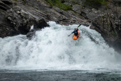 Kayaker in an orange kayak paddles through a frothy rapid between rocky walls.