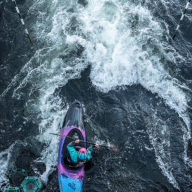 Top-down view of a kayaker navigating whitewater gates in a fast river, pink-to-blue kayak slicing through foamy water