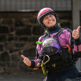 Smiling woman in a pink helmet and purple life jacket giving two thumbs up outdoors.
