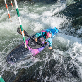 Kayaker navigating a whitewater gate in a purple kayak, wearing a pink helmet and teal jacket amid splashing water.