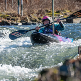 Kayaker in a pink helmet paddling through whitewater rapids, navigating between hanging slalom gates in a colorful kayak.