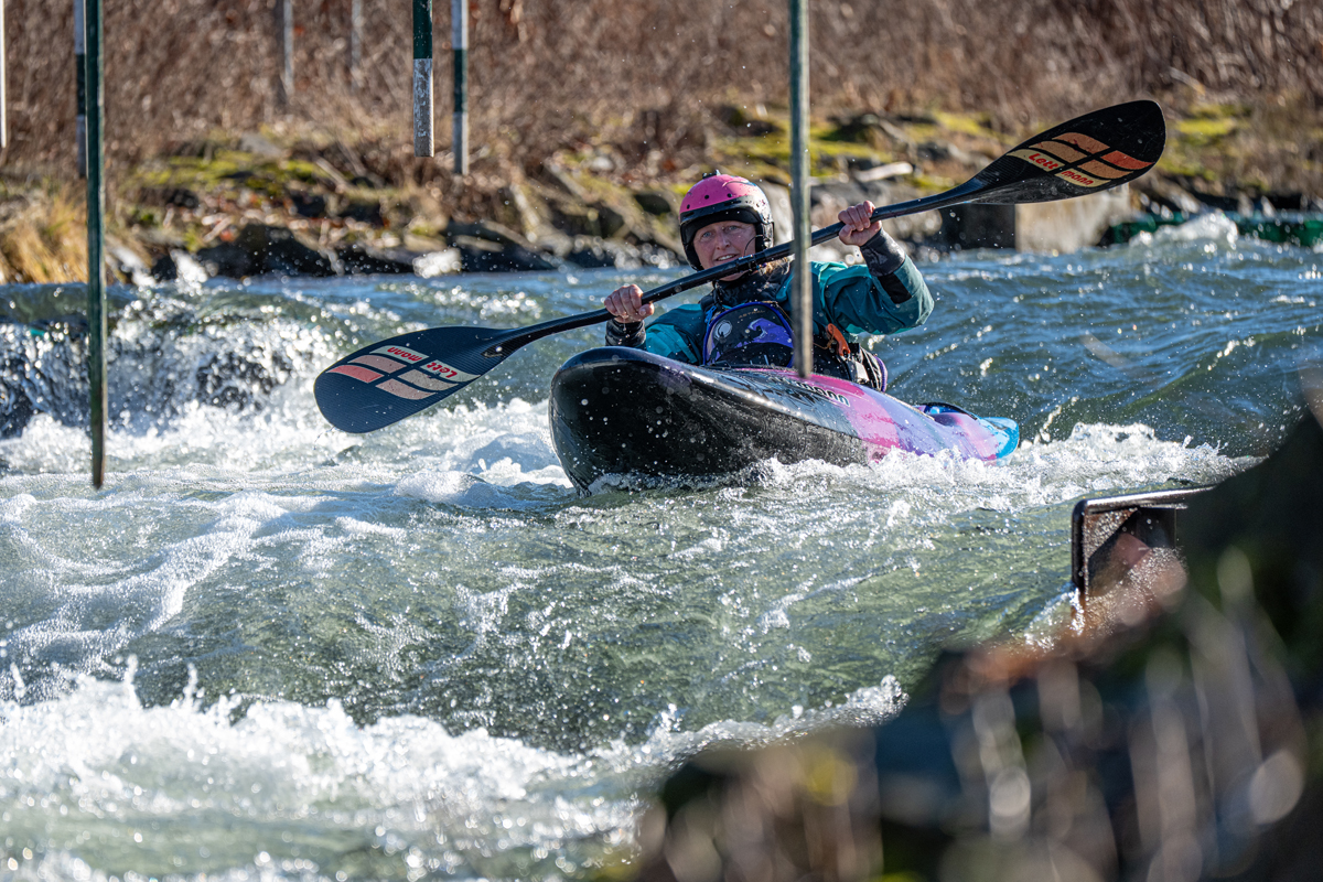 Kayaker in a pink helmet paddling through whitewater rapids, navigating between hanging slalom gates in a colorful kayak.