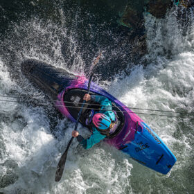 A kayaker wearing a teal jacket and pink helmet paddling a blue‑pink kayak through whitewater rapids, viewed from above.