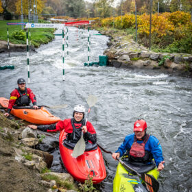 Three kayakers in bright boats navigate a whitewater slalom course with green-and-white gates on a fast-flowing river, autumn trees in the background.