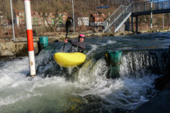 Person in a yellow kayak paddles through whitewater by a dam, wearing a pink helmet and black wetsuit.