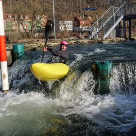 Person in a yellow kayak paddles through whitewater by a dam, wearing a pink helmet and black wetsuit.
