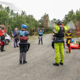 Group of kayakers in protective gear forming a circle on a gravel road, preparing for a river trip with paddles and inflatable boats nearby.