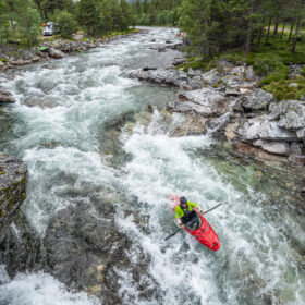 Kayaker in a bright red kayak paddling through whitewater rapids between rocky banks and pine trees.