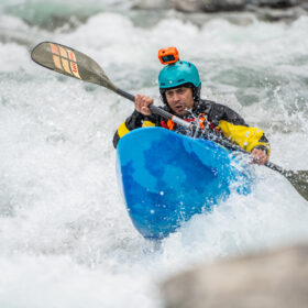 Kayaker in a blue boat paddling through whitewater rapids, wearing a teal helmet and yellow-black jacket with a mounted action camera on the helmet.