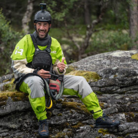 Man in neon green outdoor gear and helmet sits on a mossy rock, camera mounted on helmet, smiling at the camera.