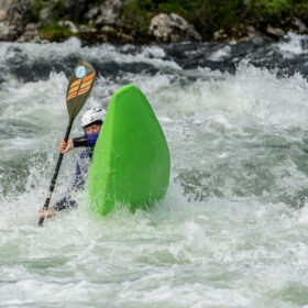 Kayaker wearing a helmet grips a paddle while leaning against a bright green kayak in turbulent whitewater.