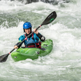 Kayaker in a blue helmet paddling a bright green kayak through whitewater rapids.