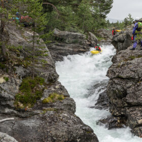 Group of kayakers navigate a whitewater rapid through a rocky canyon, with observers on the cliffs above the water. (informative)