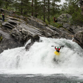 Kayaker in a yellow boat and red jacket rides a big whitewater drop through a rocky, forested gorge, spectators watching from the rocks above.