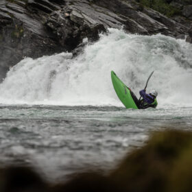 Person in a bright green kayak flips over in rough whitewater near a rocky riverbank, wearing a helmet and life jacket.
