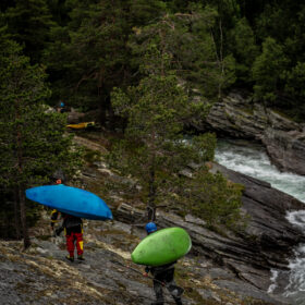 Two kayakers carry bright blue and green kayaks down a rocky slope toward a white-water river, wearing helmets and outdoor gear