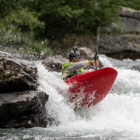 Kayaker in a red boat soloing through rough whitewater beside rocky riverbank, spray splashing around the bow.
