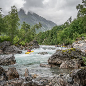 Yellow kayak navigates turquoise river rapids among large rocks with green trees and a misty mountain in the background.