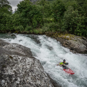 Kayaker in a bright green shirt paddling a red kayak through white-water rapids beside rocky riverbank with green forest and mountains in the background