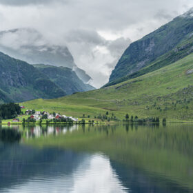 Scenic valley with a calm lake reflecting green fields, a small village of white and red-roofed houses at the shore, and towering misty mountains.