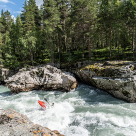 Person in a red kayak paddling through whitewater rapids between rocky cliffs with a pine forest and blue sky behind, showing an outdoor river scene on a sunny day.