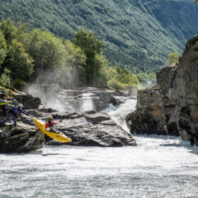 Group of kayakers in colorful gear on rocky riverbank beside a white-water rapid, surrounded by lush green forested canyon.
