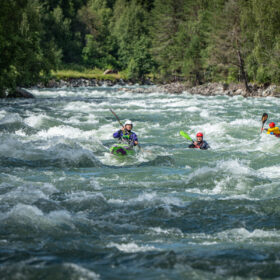 Three kayakers navigate fast whitewater through a forested river, wearing helmets and life jackets.