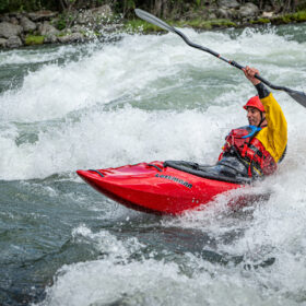 Kayaker in a red kayak rounds whitewater rapids, wearing a red helmet and yellow jacket, paddle raised overhead.