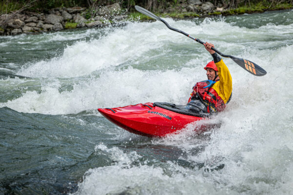 Kayaker in a red kayak rounds whitewater rapids, wearing a red helmet and yellow jacket, paddle raised overhead.