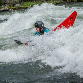 Whitewater kayaker in a teal jacket and black helmet paddles through splashing rapids; a bright red kayak rises behind them.