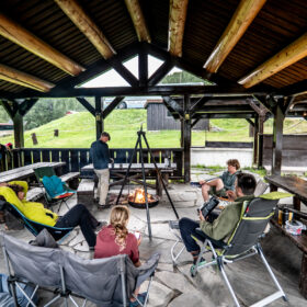 Group of people seated in folding chairs around a campfire under a wooden shelter in a park. A tripod stands near the fire as others relax at a shelter-lined pavilion.
