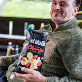 Smiling man in a green fleece jacket sits outdoors, holding a bag of El Valle Chorizo potato chips toward the camera.