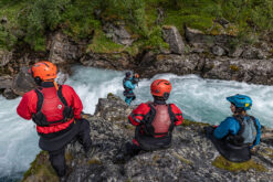 Group of four climbers/adventure participants wearing helmets and life jackets sit on a rocky ledge above a fast turquoise river in a forested gorge.