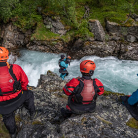 Group of four climbers/adventure participants wearing helmets and life jackets sit on a rocky ledge above a fast turquoise river in a forested gorge.