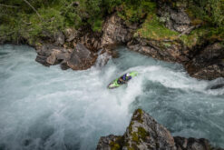 Kayaker in a bright green kayak steers through a whitewater rapid between mossy rocks near a canyon wall.