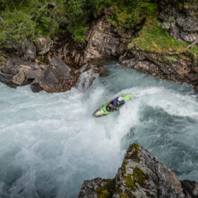 Kayaker in a bright green kayak steers through a whitewater rapid between mossy rocks near a canyon wall.
