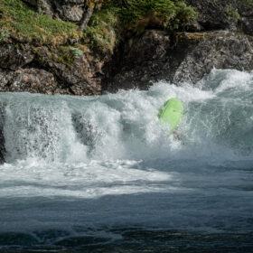 Bright neon-green kayak rides a foamy river wave near a rocky riverbank.
