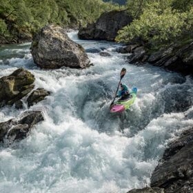 Kayaker in a colorful kayak paddles through whitewater rapids in a rocky river gorge surrounded by trees and mountains.