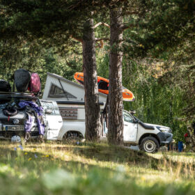 Wooded campsite with two white vans and gear piled on and around; an orange kayak rests on top between them.