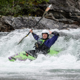 Kayaker wearing a white helmet and blue jacket navigates rough whitewater in a rocky river, green-purple kayak slicing through waves.