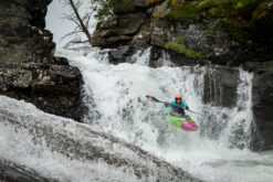 Kayaker in a bright green-pink kayak descends a waterfall between rocky cliffs with a teal wetsuit and orange helmet.
