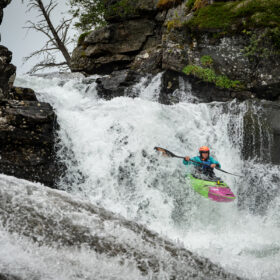 Kayaker in a bright green-pink kayak descends a waterfall between rocky cliffs with a teal wetsuit and orange helmet.