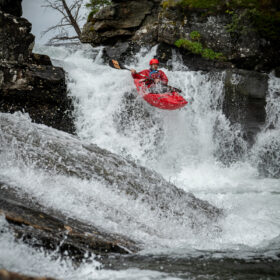 Person in a red kayak navigates whitewater over a rocky rapid between mossy cliffs, spray splashing around.
