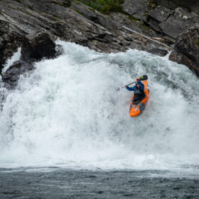 Kayaker in an orange boat rides a wave over a rocky waterfall, spray flying around him in turbulent water.