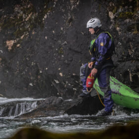 Person wearing a purple waterproof suit and white helmet stands on a rock at the edge of a fast-moving river with a green kayak nearby, ready for instruction or rescue.