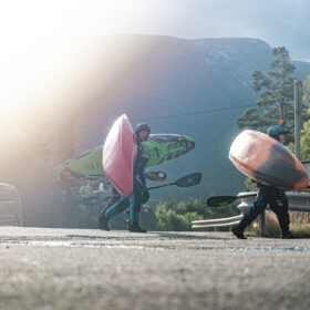 Two people wearing helmets carry large water-sports boards (kayaks) across a mountain road as a car passes by and forested hills loom in the background.