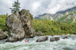 Kayaker in a red kayak paddling through whitewater near a towering rock formation at the riverbank, with forest and mountains in the background.