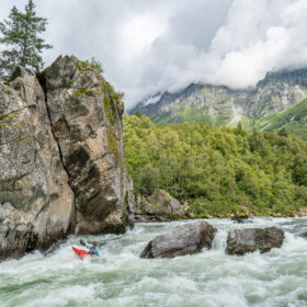 Kayaker in a red kayak paddling through whitewater near a towering rock formation at the riverbank, with forest and mountains in the background.