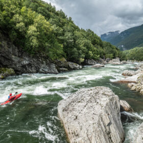 Person in a bright red kayak paddles through whitewater on a rocky, forested river canyon in a mountainous landscape.
