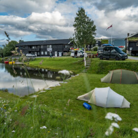 Tents pitched on a grassy hill beside a calm lake, with wildflowers in the foreground and dark wooden buildings and parked vehicles in the background under a cloudy sky.